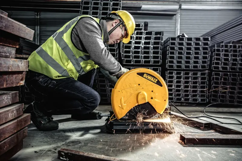 Une personne portant un équipement de sécurité utilise une scie à métaux DEWALT, modèle D28730, pour couper des barres métalliques dans un environnement industriel. Des étincelles jaillissent de la zone de coupe.