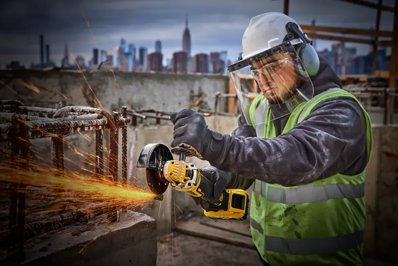 Ouvrier utilise une meuleuse DEWALT DCG405P2 sur chantier, équipement de sécurité, ville en arrière-plan.