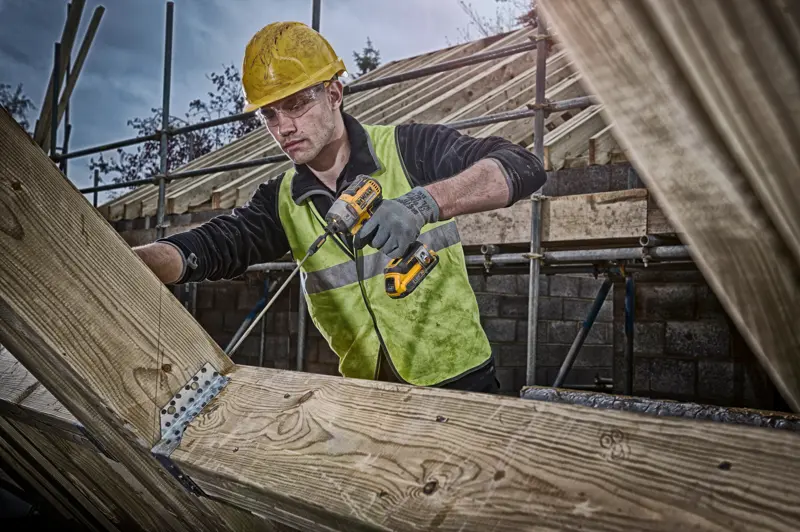 Personne utilisant une perceuse DEWALT sur des poutres en bois, avec un gilet haute visibilité et un casque de sécurité.