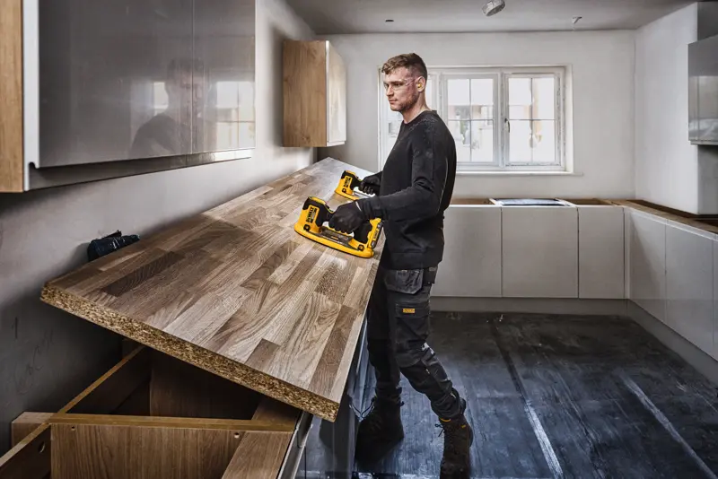 Une personne au visage flouté installe un plan de travail en bois dans une cuisine à l'aide d'un outil électrique DEWALT. La cuisine est moderne avec des armoires de couleur claire et une grande fenêtre à l'arrière-plan.