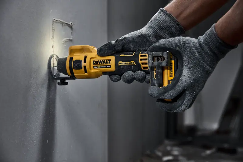 Outil rotatif sans fil DEWALT découpant du placoplâtre, tenu par des mains gantées.