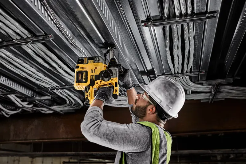 Une personne portant un casque, des lunettes de sécurité, des gants et un gilet de sécurité utilise une perceuse rotative sans fil jaune DEWALT pour percer un plafond métallique avec des câbles électriques.
