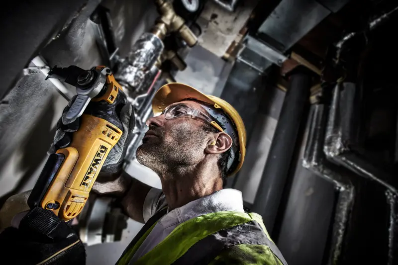 Ouvrier coupe un tuyau mural avec un DEWALT DWE315, équipé d'un casque et d'un gilet dans un espace industriel.