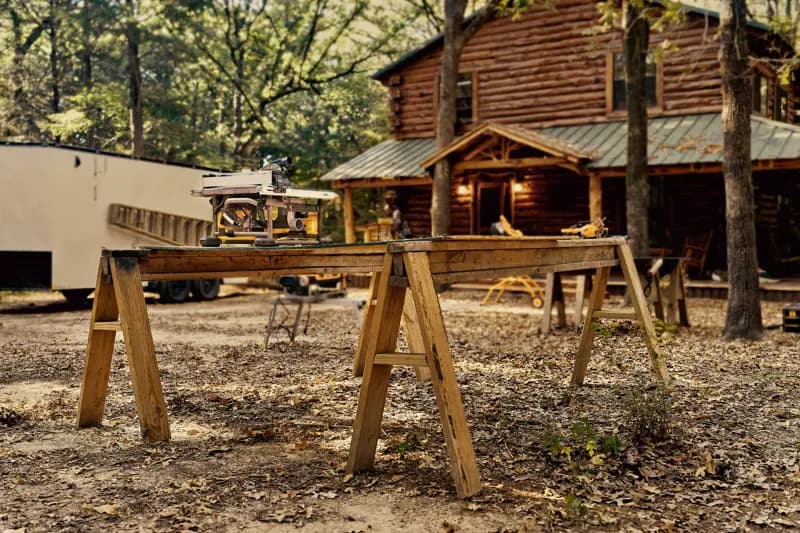 Une scie à table DEWALT installée sur un établi en bois composé de deux tréteaux à l'extérieur, avec une cabane en rondins et des arbres en arrière-plan.
