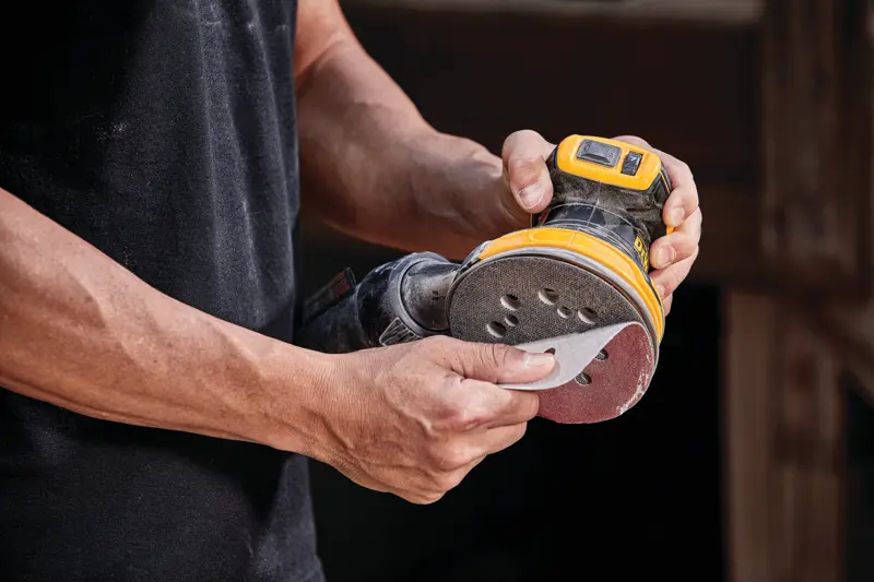 Une personne tient une ponceuse orbitale aléatoire sans fil DEWALT, modèle DCW210B, et fixe un disque de ponçage sur l’outil.