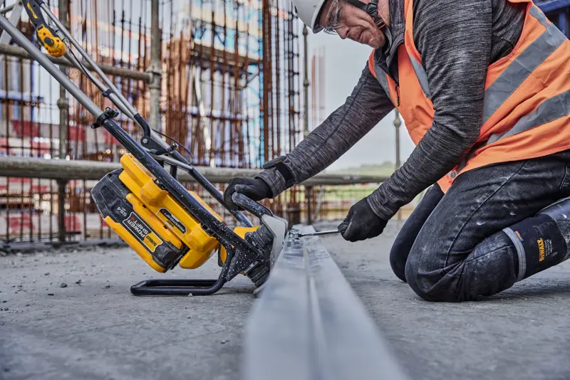 Ouvrier utilise un outil jaune DEWALT pour scier du métal sur chantier, en gilet orange et équipement de sécurité.