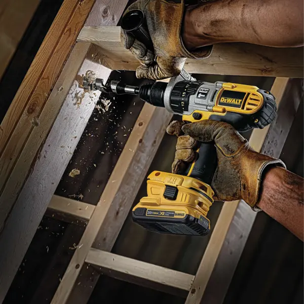 Une personne portant des gants de travail marron utilise une perceuse électrique sans fil DEWALT pour percer un trou dans une poutre en bois, avec des copeaux de bois projetés par le processus de perçage.