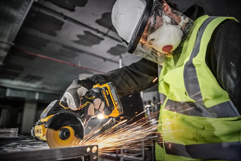Une personne portant des équipements de sécurité, dont un casque, une visière, un masque, des gants et un gilet haute visibilité, utilise un outil électrique sans fil DEWALT DCS438N pour couper du métal, générant des étincelles.
