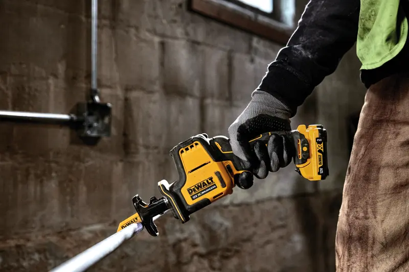 Main gantée tenant une scie sabre sans fil DEWALT DCS312D2 coupant un tuyau métallique en intérieur.