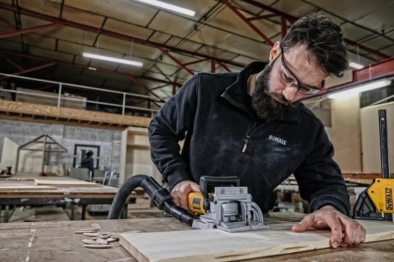 Une personne utilise un outil de menuiserie DEWALT DW682K pour couper une planche de bois sur une table dans un atelier. Le visage de la personne est masqué pour préserver sa confidentialité.