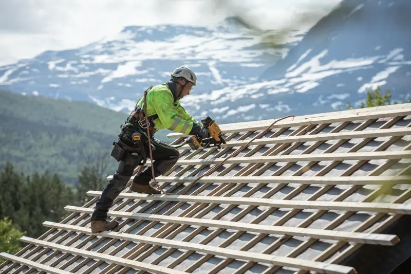 Ouvrier utilisant une cloueuse DEWALT sur un toit incliné, équipé de sécurité, montagnes en arrière-plan.