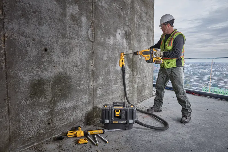 Un ouvrier du bâtiment portant un gilet de sécurité et un casque utilise une perceuse sans fil DEWALT avec un accessoire d’extraction de poussière pour percer un mur en béton. L’ouvrier se tient sur un grand bâtiment avec une vue sur la ville à l’arrière-plan. Plusieurs outils DEWALT et une mallette à outils sont posés au sol à proximité.