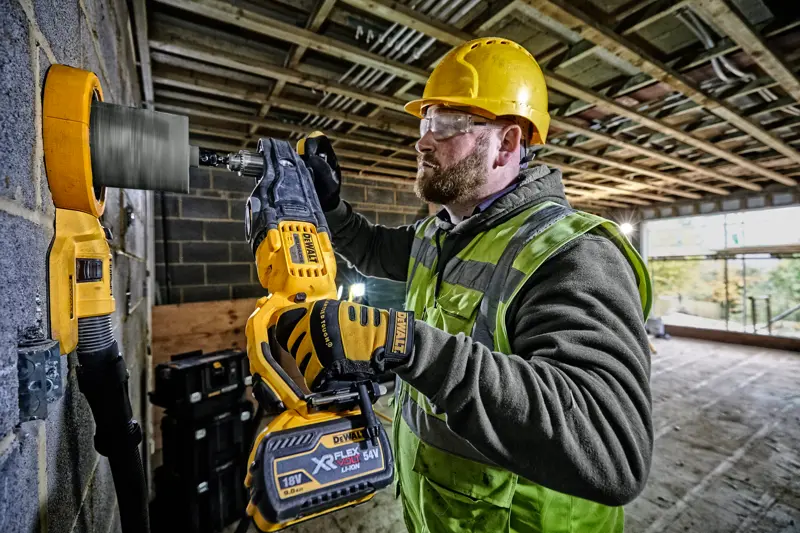 Un ouvrier du bâtiment portant un casque de sécurité jaune et un gilet haute visibilité utilise une perceuse sans fil DEWALT DCD470N avec une scie-cloche pour percer un mur en béton sur un chantier.