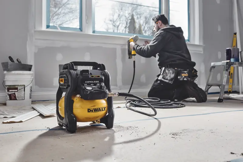 Compresseur sans fil DEWALT dans une pièce rénovée, ouvrier utilisant un cloueur près d'une fenêtre.