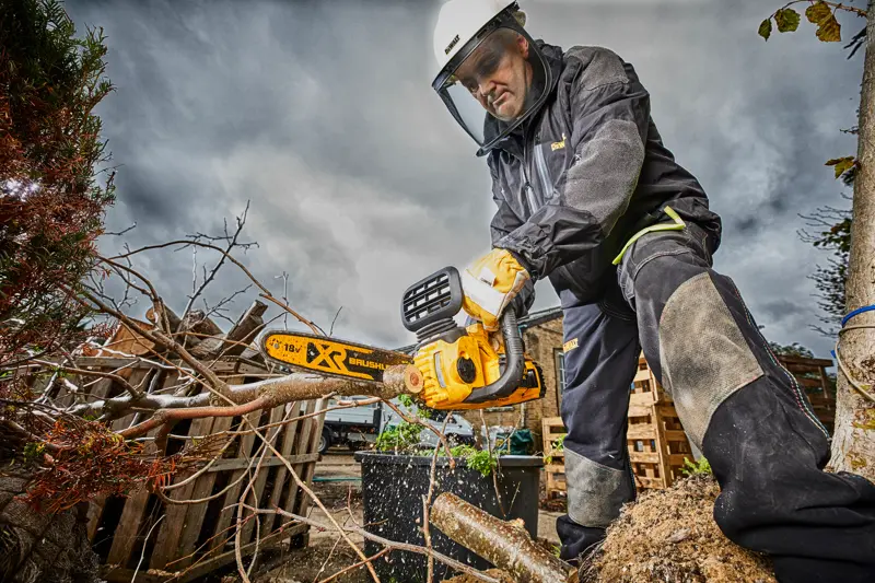 Personne en tenue de sécurité coupe des branches avec une tronçonneuse DEWALT dehors par temps nuageux.