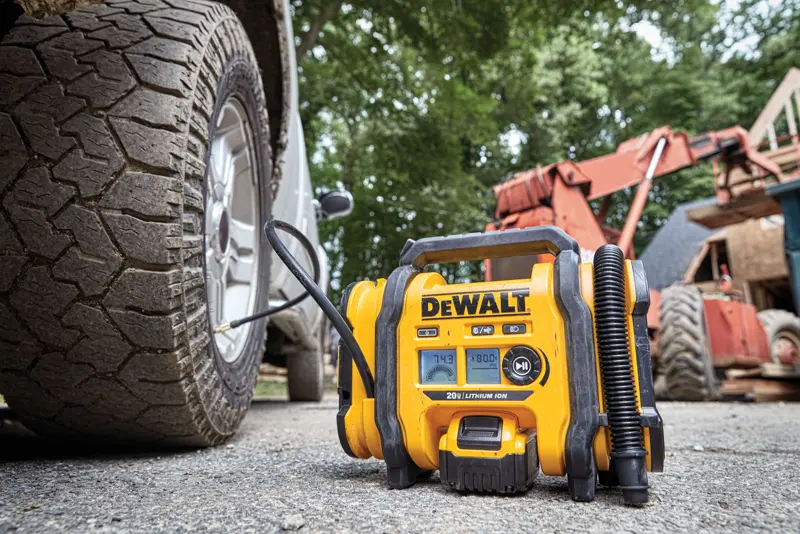 Un gonfleur de pneus portable DEWALT est montré en utilisation, connecté à un pneu de véhicule, sur un chantier extérieur avec des équipements de construction en arrière-plan.
