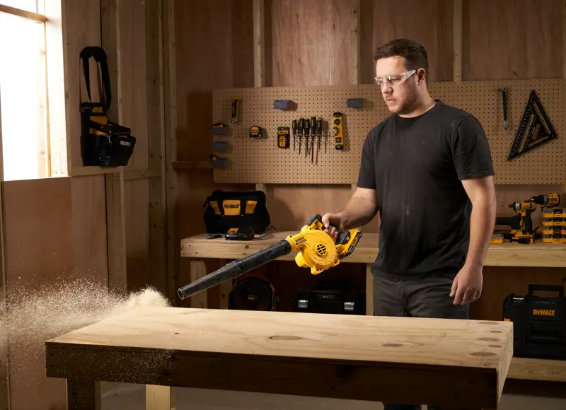 Personne utilisant un souffleur DEWALT DCV100 pour nettoyer la sciure d’un établi en bois dans un atelier.