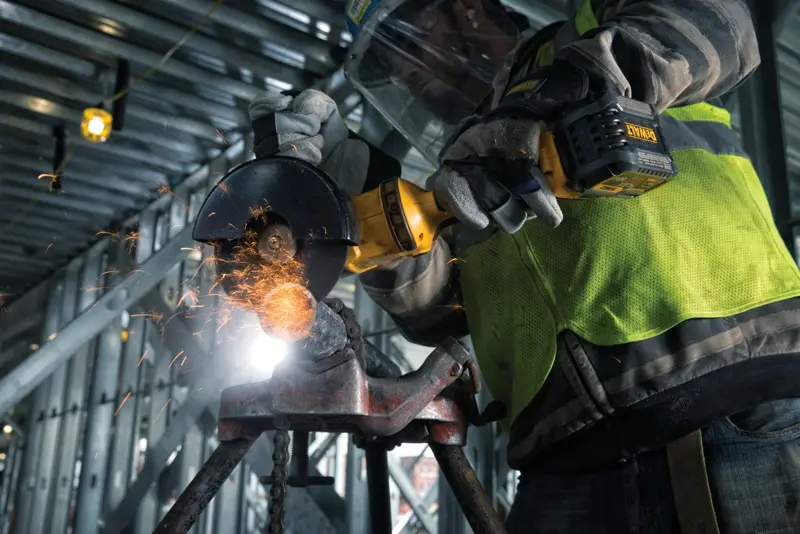 Gros plan sur une personne portant des gants de protection et un gilet de sécurité utilisant une meuleuse angulaire sans fil DEWALT pour couper du métal, avec des étincelles, sur un chantier industriel.